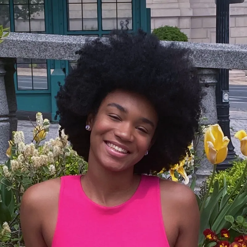 Student Sienna Jade A'isha Miller, a class of 2025 Social Work major, sits in front of a street garden in center city Philadelphia. She is wearing a pink sleeveless top and smiling broadly.
