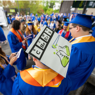 Students preparing for commencement, in blue robes and mortarboards. The central student's mortarboard has been decorated with chemical symbols reading "Cheers."