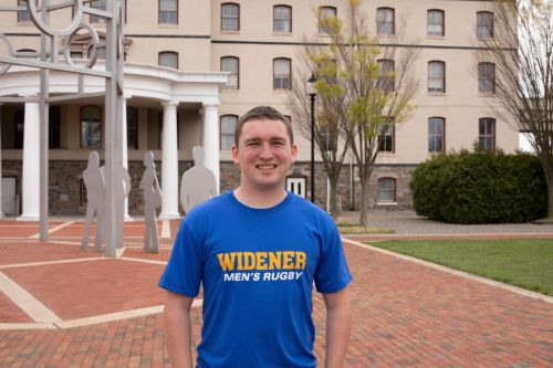 Michael Fehrle from the Widener class of 2022 stands in front of the Bown Garden, wearing a blue Widener Rugby t-shirt