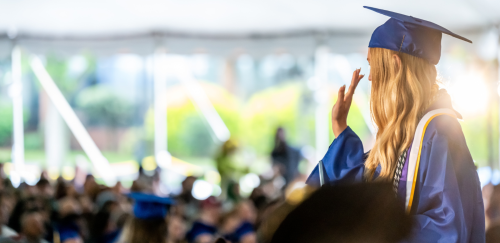 A Widener grad stands and waves. She is wearing a blue mortarboard and blue graduation robes. In the distance is a sea of other graduates.