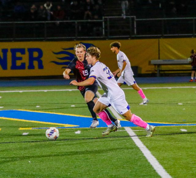 A Widener soccer player advances the ball against a player from Arcadia. Widener players are wearing white shirts and shorts with pink socks, and the Arcadia players are wearing black shirts, shorts, and socks.